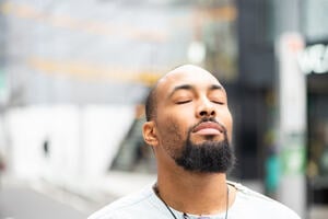 A man with beard standing on the street with his eyes closed, enjoying a moment of peace.