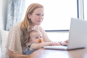 A mother works on her computer while holding her infant baby in her lap.