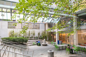 An interior courtyard of a building is shown. The building is concrete and glass, with tables in the courtyard with green chairs. There is a tree overhead and greenery throughout the courtyard