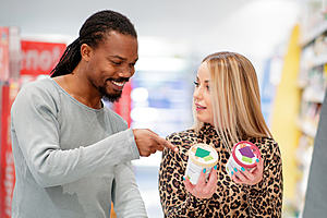 A man and a woman are choosing between two tubs of icecream in a supermarket. The man is pointing at the label of one and the woman is looking at him questioningly.
