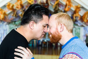 Two men stand in profile, pressing their noses together in a traditional Māori hongi. The man on the left is Māori with dark hair and brown skin. The man on the right is European with light skin and ginger hair and beard. They stand in front of a marae.