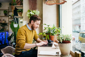 A young man sitting in a small café filled with green plants, using his laptop writing.