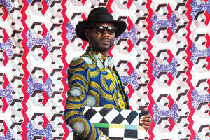 A man poses dressed in a colorfully patterned suit with a large hat, sunglasses and bag.