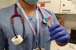 A medical professional wearing blue scrubs, stethoscope with a white tape roll on it, a transgender flag colour lanyard and two medical tubes with the trans blue and pink colour tops on them   
