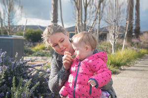 The photo is of a female cancer patient walking on a garden path holding her young infant daughter. In the photo, the patient is holding a flower up to her daughter's nose. 