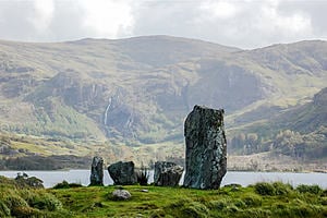 Standing stones on a grass mound set against a backdrop of mountains and a lake.