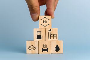 Fingers holding a wooden cube on top of a pile of five other cubes with various hydrogen related symbols.