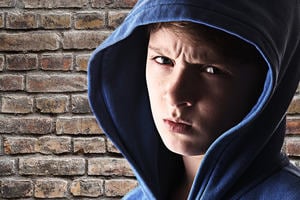 Boy in a hooded shirt scowling in front of a brick wall.