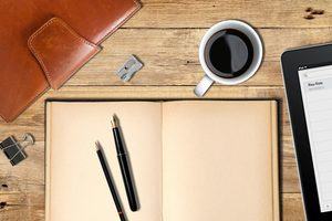 A pen and pencil sit on an open notebook with a coffee and an iPad close at hand, ready to start writing fiction