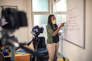 A teacher with a face mask on, standing at a full size whiteboard, underlining sentences with a dry marker. In the foreground are two cameras which she is presenting to