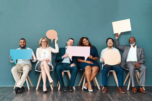 Shot portrait of a diverse group of business people with men and women holding up speech bubbles while they wait in line in front of a blue wall.