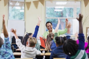 Teacher smiling at a group of primary age students with their hands up