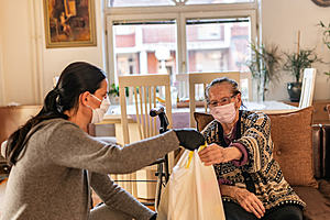 Two women wearing face masks sat down. The younger woman is handing the older woman a plastic bag
