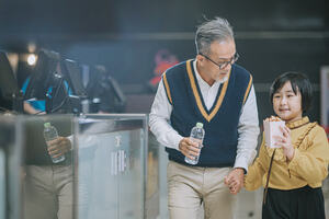 A Chinese grandfather holding his granddaughters hand, he is also holding a bottle of water and she holding a cup of popcorn. They are in conversation as they are entering the movie theatre, having just brought movie tickets