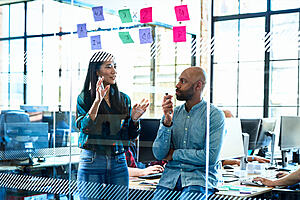 Two people talking in front of a glass wall with post it notes on it.