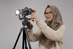 An image of a woman wearing a head scarf and jumper. She is adjusting a smartphone set up for filming atop a tripod.